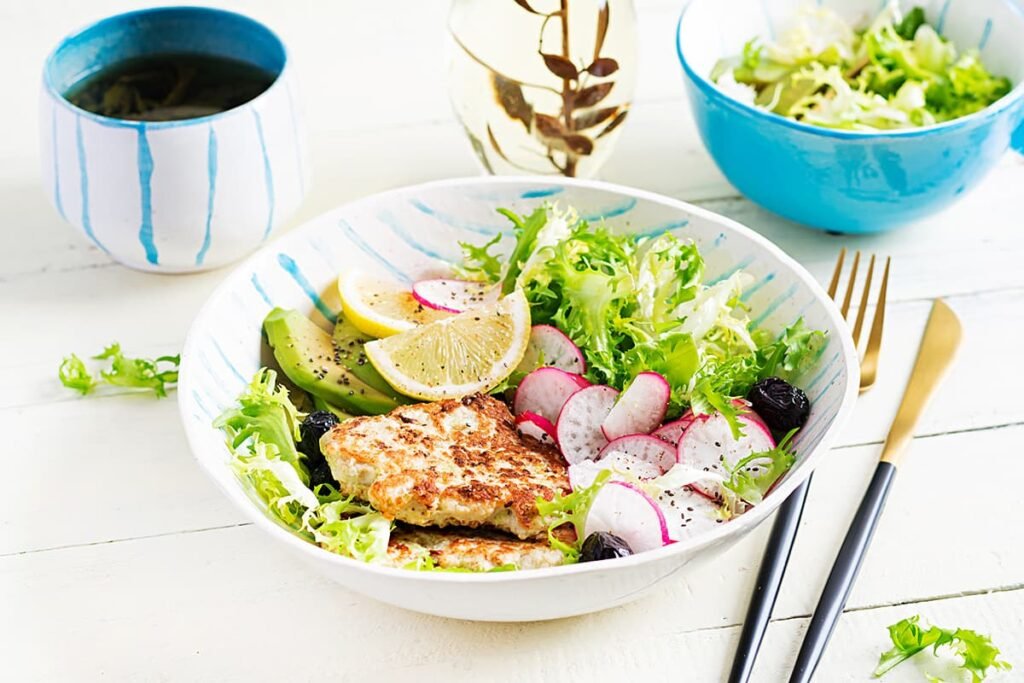 Kipburger geserveerd in ene bowl met sla, avocado, radijs en olijven